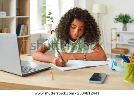 African American teenage girl handwriting in notebook while doing homework using laptop. Concentrated cute curly girl sitting at table in her room and studying remotely. Homeschooling concept. Royalty-Free Stock Photo #2304812441