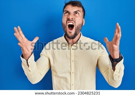 Handsome hispanic man standing over blue background crazy and mad shouting and yelling with aggressive expression and arms raised. frustration concept.  Royalty-Free Stock Photo #2304825231