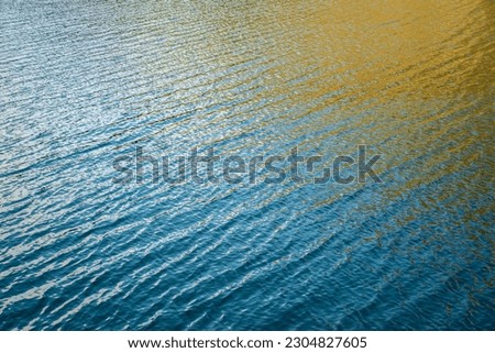 Wavy surface of a lake with blue water and a contrasting yellow reflection on it. Picture taken from above the surface of the lake, located in the Alps mountains in Northern Italy. 