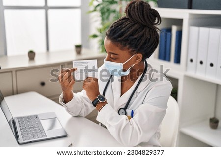 African american woman doctor wearing medical mask holding covid-19 vaccination record card at clinic