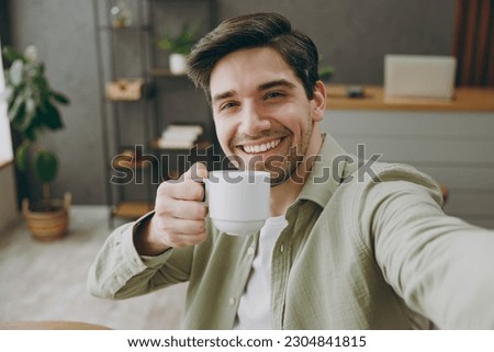 Close up fun young man wear shirt casual clothes doing selfie shot pov on mobile cell phone drink tea sitting alone at table in coffee shop cafe restaurant indoors rest relax during free time inside