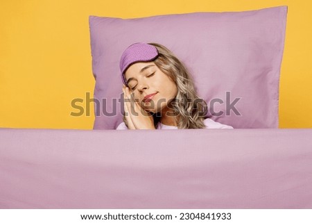 Young calm woman wear purple pyjamas jam sleep eye mask rest relax at home lies wrap covered with blanket duvet hand under cheek isolated on plain yellow background studio. Good mood night nap concept