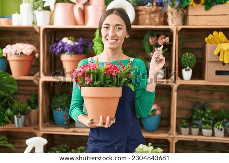 Young hispanic woman working at florist shop holding plant smiling happy pointing with hand and finger to the side 