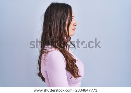 Young brunette woman standing over blue background looking to side, relax profile pose with natural face and confident smile.  Royalty-Free Stock Photo #2304847771