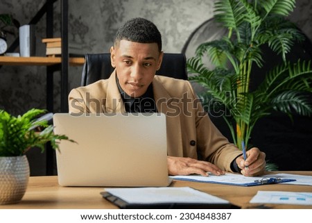 A young black man sits in the office and carefully looks at the laptop screen. Male businessman sitting at home office signing documents.