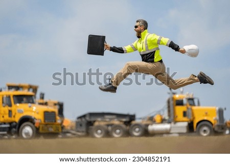 Hispanic 40 s builder excited jump on site construction. Excited builder construction worker in a safety helmet jumping in front of the trucks. Excited crazy builder man in helmet jump outdoor. Royalty-Free Stock Photo #2304852191