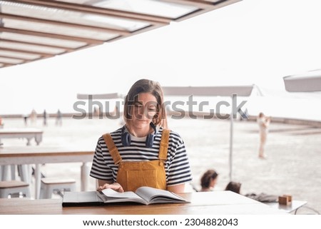 Young woman reading a book in a cafe in yellow dungarees outdoors. Concept: lifestyle, fashion style, outdoors.
