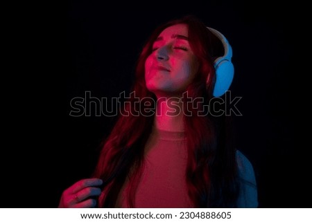 Fashion hipster girl smiles and wear headphones listening to music over color neon background at studio. Dancing in modern wireless headphones. Colored gel portraits