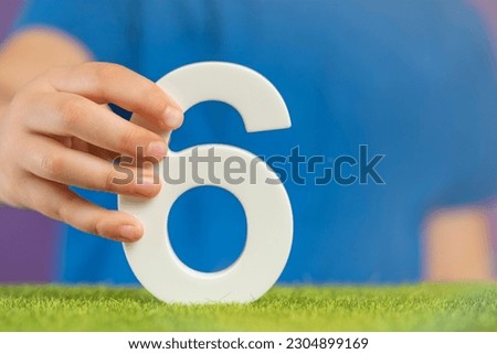 Number 6 in hand. A hand holds a white number six on green grass and a purple background, six percent per annum. Swedish Constitution Day. July 6th. Royalty-Free Stock Photo #2304899169