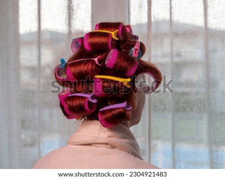 Rear view of a young woman with hair curlers posing against a light curtain background at home
