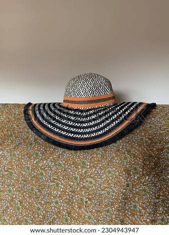 Woven brightly coloured women's summer hat for the beach with black and orange stripes. Lays on a fine floral print fabric, in daylight, weave texture.