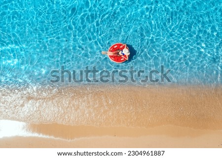 Aerial view of a woman in hat swimming with red swim ring in blue sea at sunrise in summer. Tropical landscape with girl, clear water, waves, sandy beach. Top view. Vacation. Sardinia island, Italy	 Royalty-Free Stock Photo #2304961887