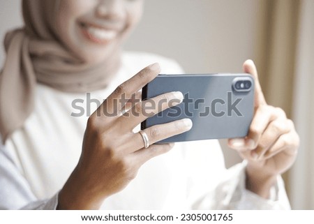 A beautiful Asian Muslim girl in casual clothes is using a smartphone taking pictures and smiling while standing by the window