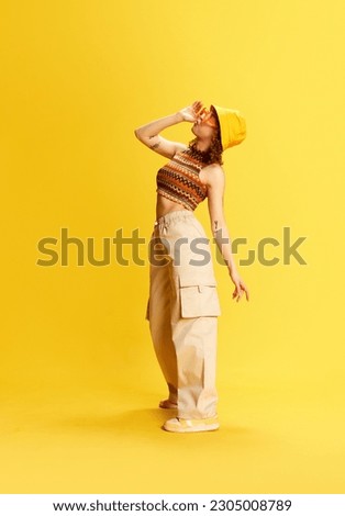 Full-length portrait of stylish young girl in trendy cargo pants, knitted top and panama standing against yellow studio background. Concept of human emotions, youth culture, fashion, lifestyle Royalty-Free Stock Photo #2305008789