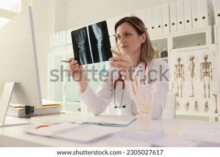 Female doctor studies X-ray picture of patient hand and arm sitting at desk in clinic office. Traumatologist controls rehabilitation process after injury