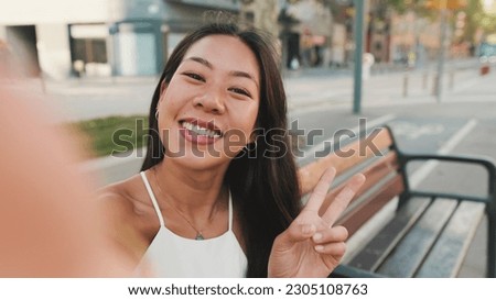 Pretty brunette girl dressed in white top and jeans is smiling making selfie while sitting on bench on cityscape background