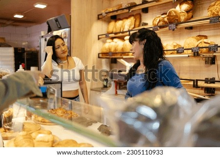 Two adult sales assistants women with casual clothes talking to each other while a client pays with credit card in a bakery