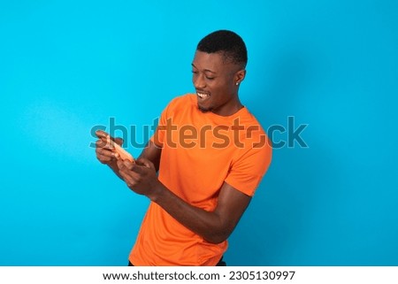 young man wearing orange T-shirt over blue studio background holding in hands cell playing video games or chatting