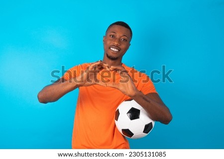 young man wearing orange T-shirt holding a ball over blue studio background smiling in love doing heart symbol shape with hands. Romantic concept.