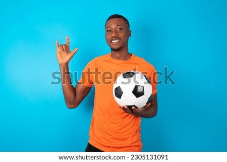 young man wearing orange T-shirt holding a ball over blue studio background doing a rock gesture and smiling to the camera. Ready to go to her favorite band concert.