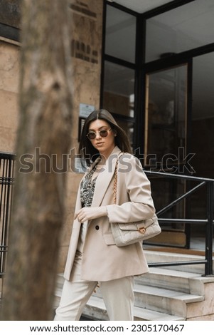 young brunette woman in stylish sunglasses and trendy outfit with white pants and beige blazer walking with handbag near modern building and blurred tree trunk on street in Istanbul Royalty-Free Stock Photo #2305170467
