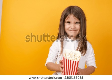 Beautiful little girl wearing white T-shirt over yellow studio background eating popcorn while watching her favourite TV show. 