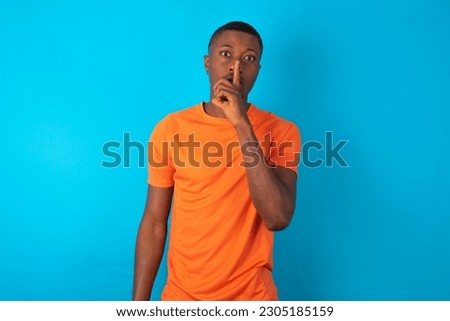 young man wearing orange T-shirt over blue studio background makes silence gesture, keeps finger over lips. Silence and secret concept.