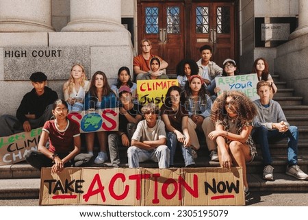 Group of climate change activists looking at the camera while sitting outside a high court with posters and banners. Multicultural young people protesting against global warming and pollution. Royalty-Free Stock Photo #2305195079