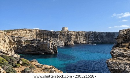 Teal blue waters shimmer, embraced by cliffs that stand tall on either side of the frame and the St. Mary's historic fortified stone watchtower is seen in the distance. Royalty-Free Stock Photo #2305215187