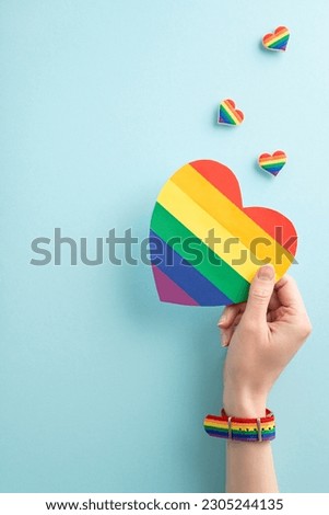 Overhead first person view vertical shot of a young woman's hand wearing a symbolic wristlet, holding a pride flag card over badges on a pastel blue background with empty space for a message or ad Royalty-Free Stock Photo #2305244135