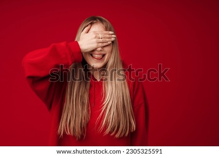 Young blonde woman showing her tongue while covering her eyes isolated over red background