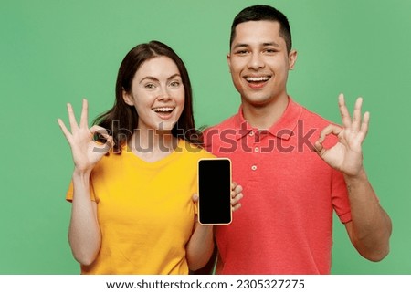 Young smiling fun couple two friends family man woman wear basic t-shirts together hold use mobile cell phone blank screen workspace area show ok isolated on pastel plain light green color background