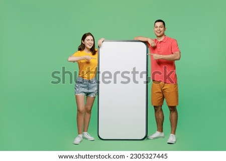 Full body smiling fun young couple two friends family man woman wear t-shirts together point finger on big huge blank screen mobile cell phone with area isolated on pastel plain light green background