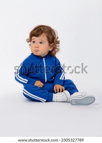 An offended kid 1-2 years old is going to cry with his lip rolled up. Sitting in a blue tracksuit and sneakers with his legs bent on a white background