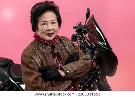 Smiling senior woman wears leather jacket with arms crossed and standing next to motorcycle over pink background, looking at camera