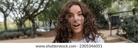 Portrait of shocked young and curly woman in white t-shirt looking at camera with open mouth while spending time in blurred park at daytime in Barcelona, Spain, banner