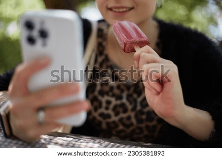 Happy young woman eating an ice cream and browsing a mobile app outdoor. Unrecognizable female person enjoying organic dessert food and using a modern smart phone in a green park