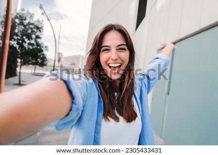 Happy young caucasian student lady looking at camera and taking a selfie portrait having fun, standing outside. Front view of laughing woman shooting a photo for social media at the university campus