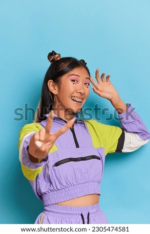 Oriental girl dressed in stylish violet-colored sportwear stands on blue studio background raised one hand to her forehead and shows victory sign with other, enjoyment concept, copy space