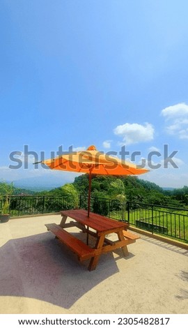 Picnic table. Table made of wood. Equipped with a bright orange umbrella. Against the background of hills and clear blue sky. Picture taken during the day.