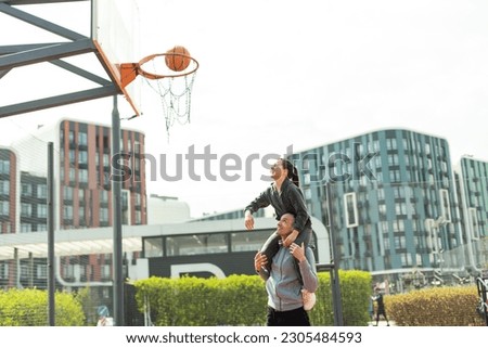 Father Carrying Daughter On Shoulders Helping Her To Put Basketball In Net
