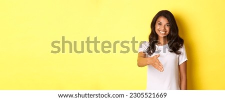 Portrait of attractive african-american girl smiling, extending hand for handshake, greeting you, standing over yellow background.