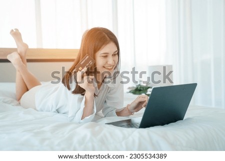 Photo of young happy woman in pajama holding credit card and talk video call Online with friends while sitting on bed.