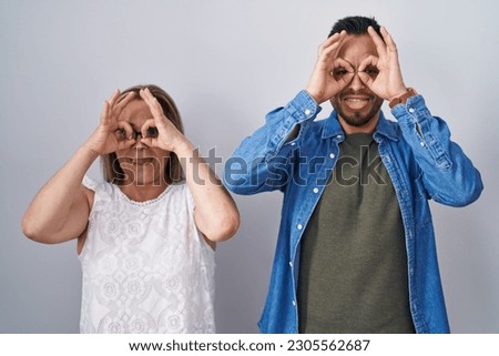 Hispanic mother and son standing together doing ok gesture like binoculars sticking tongue out, eyes looking through fingers. crazy expression. 
