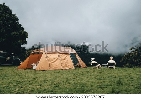 a couple sitting on wooden chairs next to a brown tent on green grass in the wild with white clouds and mountains Royalty-Free Stock Photo #2305578987