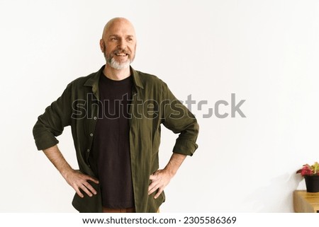 Smiling mid-aged bald man with silver beard standing confidently in front of white wall. His positive posture and expression radiate happiness and joy, showcasing contentment and cheerful demeanor Royalty-Free Stock Photo #2305586369