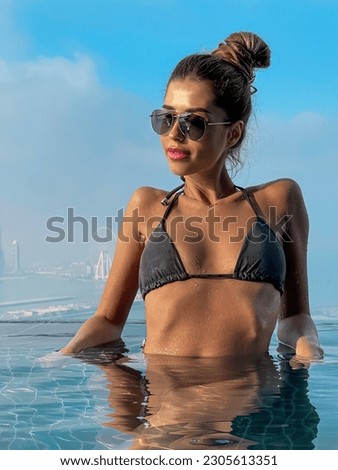 Beautiful brunette girl in a swimsuit with a slender figure, plump lips, in sunglasses. A girl in an infinite pool against the backdrop of a beautiful city. Sky clouds sea, vacation beach summer Dubai
