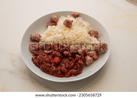a white ceramic plate with rice and red beans with sauce and pork sausages on a marble table
