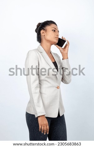 Corporate office lady with black hair having playful moment with pen, smart phone in formal wear wearing white blazer against white background. 