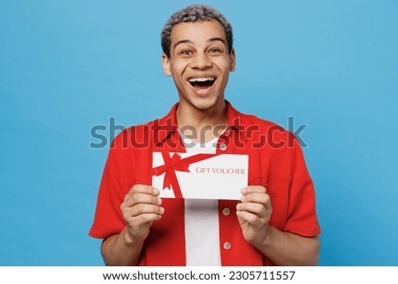 Young surprised man of African American ethnicity 20s wear red shirt hold gift certificate coupon voucher card for store isolated on plain pastel light blue cyan background. People lifestyle concept